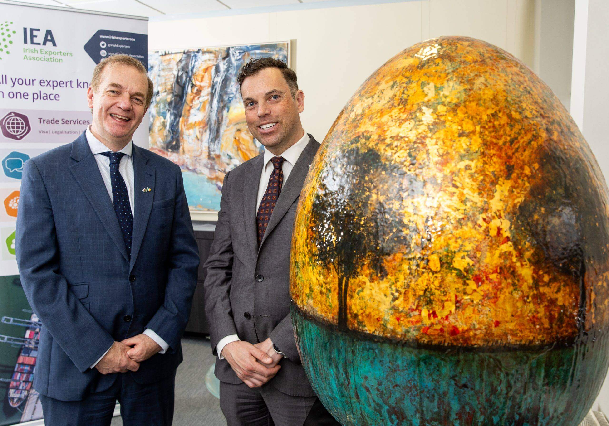 Two men in business suits stand smiling next to a large, decorative egg-shaped sculpture featuring a vibrant landscape painting, with an Irish Exporters Association banner in the background.