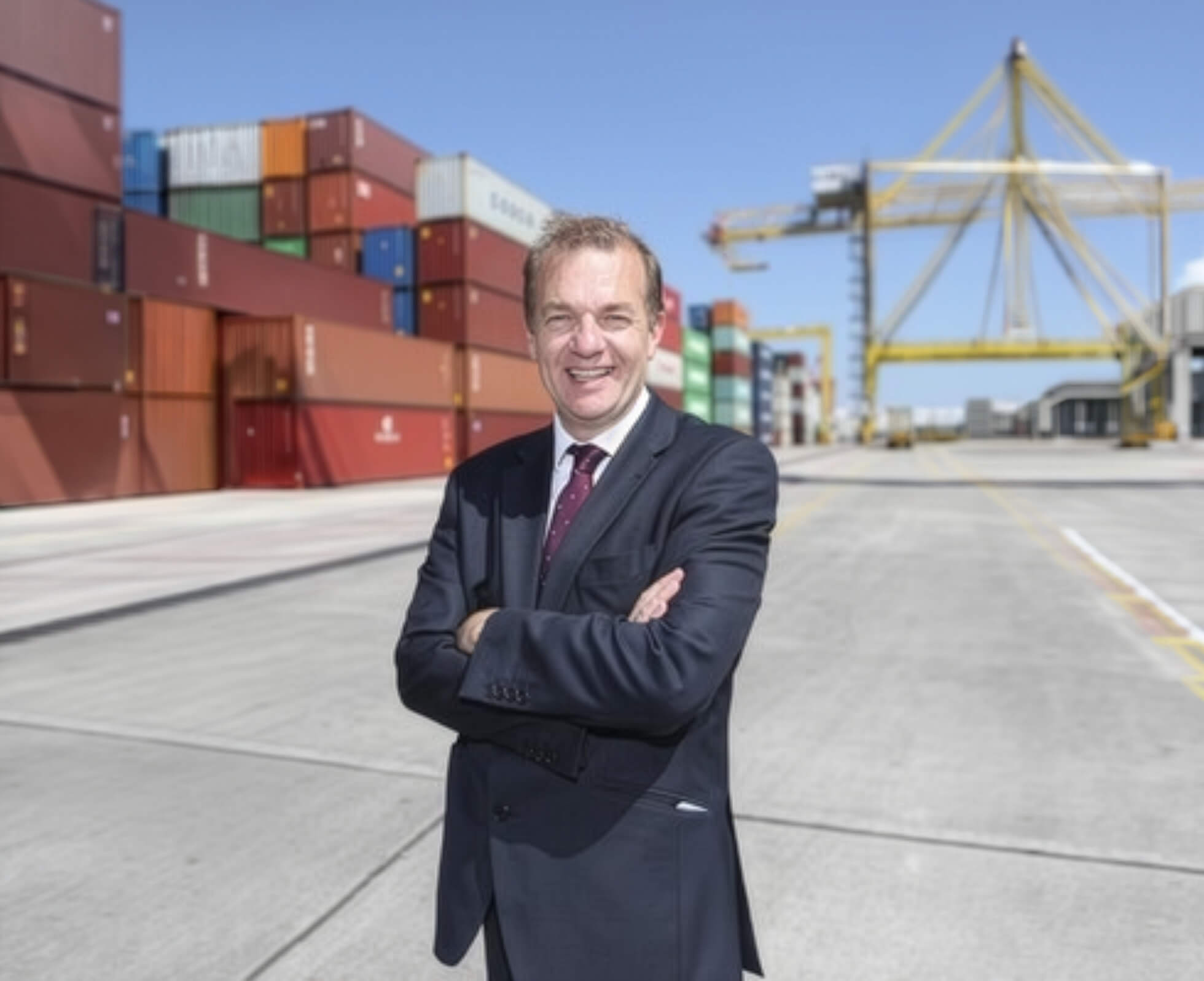 A smiling businessman in a dark suit stands with arms crossed at a shipping port, with stacks of colorful cargo containers and a yellow gantry crane in the background.